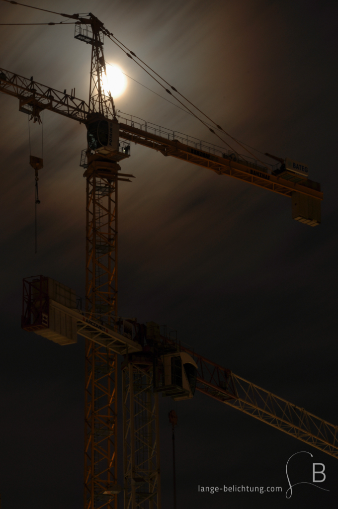 Zwei Kräne bei Vollmond auf einer Berliner Baustelle. Durch die verwischten Wolken hindurch scheint der Vollmond.