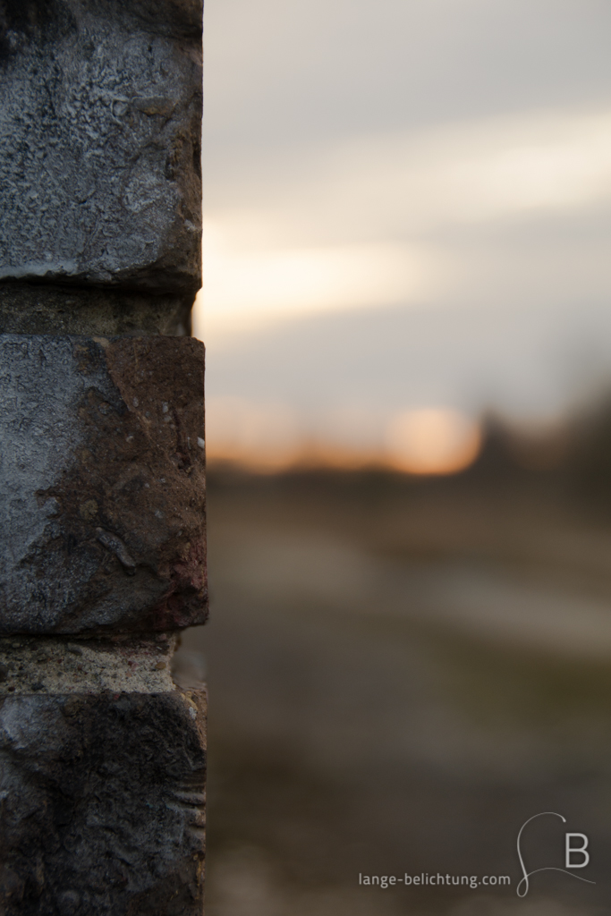 Nahaufnahme einer Backsteinmauer. Im Hintergrund sieht man eine große Freifläche. Am Horizont taucht die Sonne den Himmel in oranges Licht.