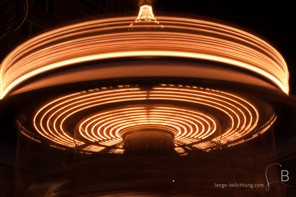 Ein historisches Karussell auf dem Weihnachtsmarkt in Leipzig. Durch die Langzeitbelichtung wird die Drehung des Karussells sichtbar. Im Hintergrund sind weitere Buden zu sehen.