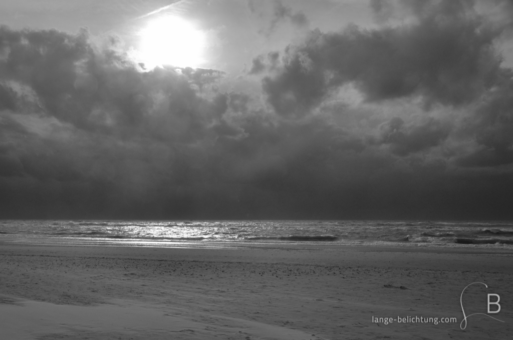 An der Nordsee Küste wird ein aufziehender Sturm von dunklen Wolken angekündigt. Der Strand ist menschenleer. Durch die Wolken bricht aber noch mal die Sonne und spiegelt sich im Wasser.