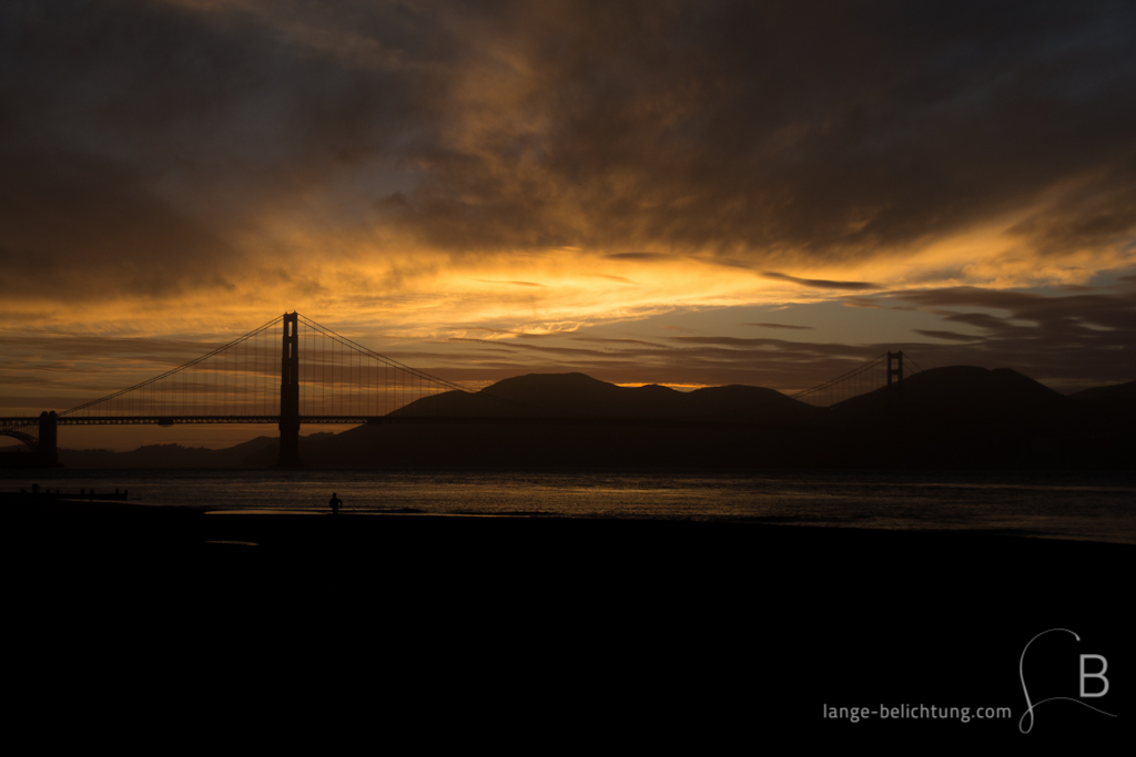 Die Silhouette der Golden Gate Bridge im Sonnenuntergang. Im Vordergrund sieht man einen Jogger am Strand laufen. Die Wolken färben den Himmel orange-rot.