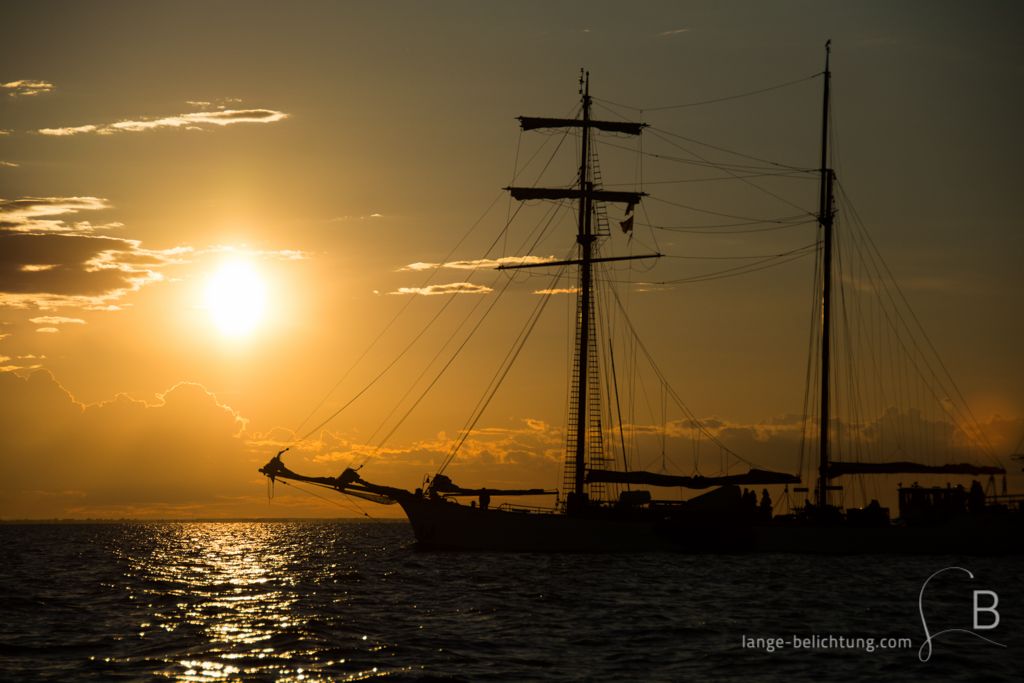 Die Sonne geht auf der Ostsee unter, während ein Zweimaster mit geborgenen Segeln als Silhouette im Vordergrund fährt. Die wenigen Wolken am Himmel sind von der Sonne orange gefärbt.