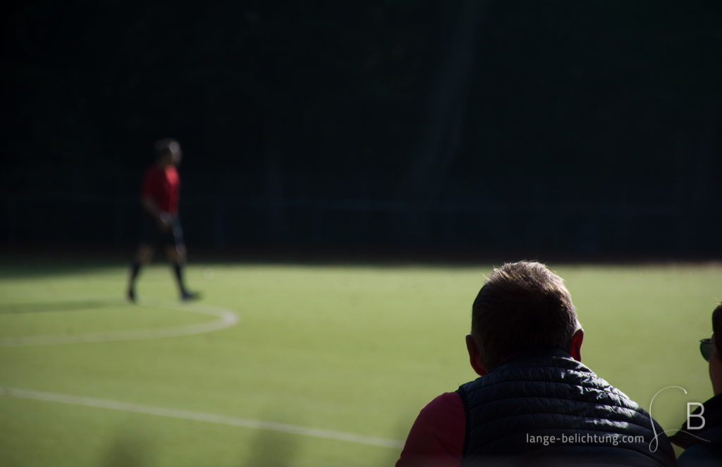 Ein Zuschauer beobachtet den Schiedrichter in rot, der allein über den Kunstrasenplatz bei einem Fußballspiel läuft.dd