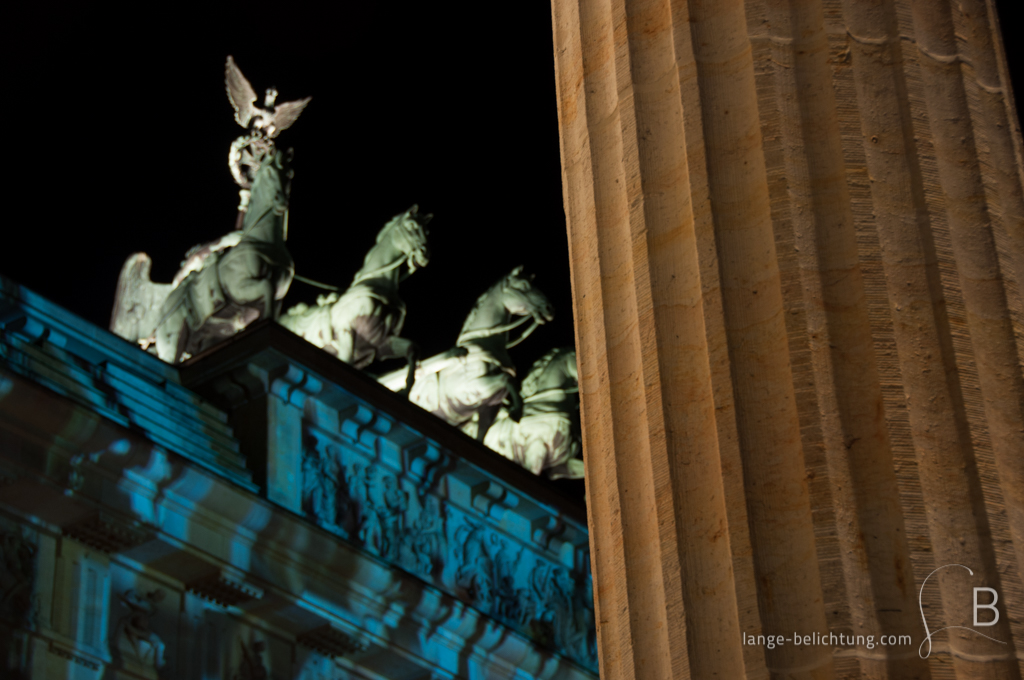 Die angeleuchtete Quadriga strehlt über dem Brandenburger Tor. Im Vordergrund des Bildes ist eine Säule des Seitenbaus am Brandenburger Tor zu sehen.