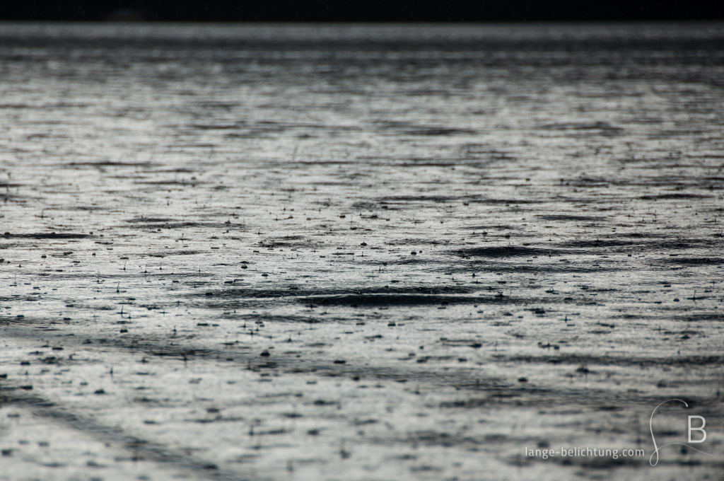 Bei einem starken Sommerregen fallen Regentropfen in den See und lassen viele kleine Wasserspritzer entstehen.