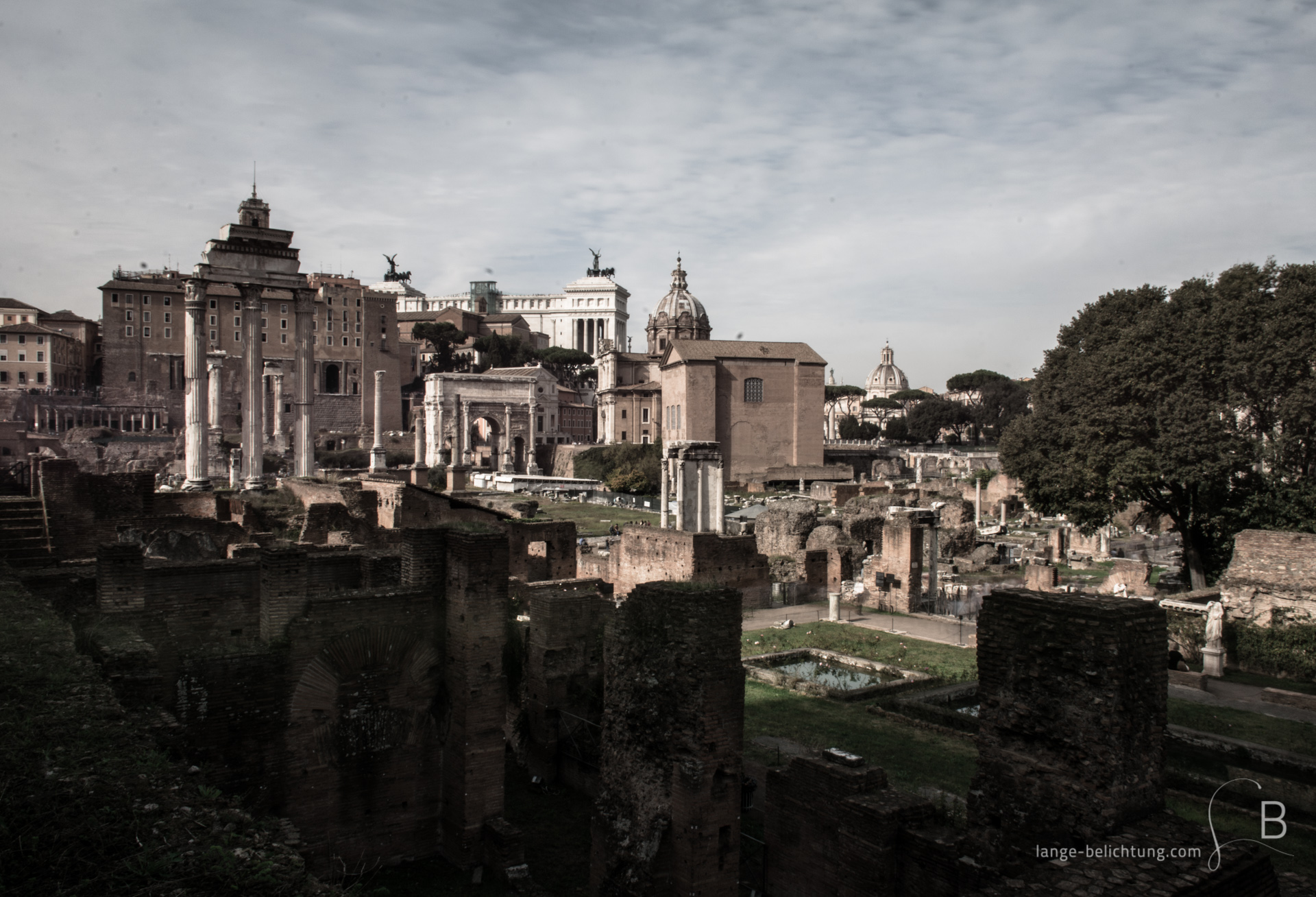 Ein Blick auf das Forum Romanum. Im Vordergrund stehen Mauerreste. Weiter hinten sieht man Überreste von Tempeln und den Triumpfbogen. Zwischen den Gebäuden erkennt man auch das Denkmal Vittorio Emmanuel.