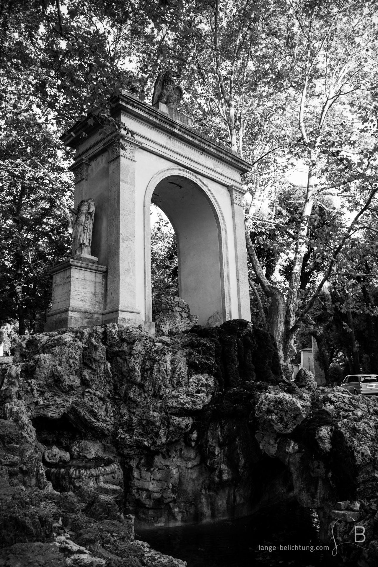 Abgebildet ist ein Brunnen in Rom nahe der Villa Borghese zwischen herbstlichen Laubbäumen. Das Bild in schwarzweiß zeigt die Fontana di Esculapio.