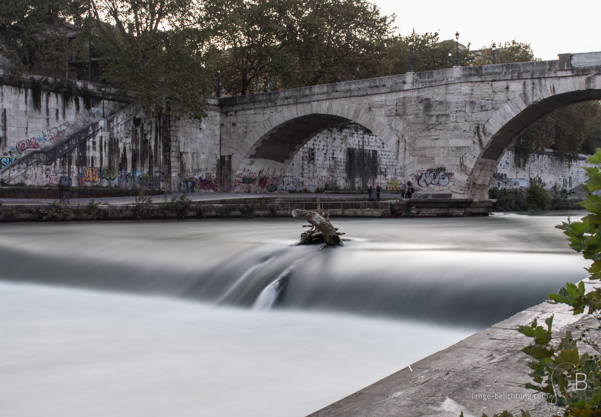 Eine Brücke, die übersäht ist mit Graffiti, führt über den Fluss Tiber zur Tiberinsel. Im Vordergrund ein kleines Wehr, dass das Wasser schneller fließen lässt. Kurz vor dem Wehr steckt ein Baumstamm